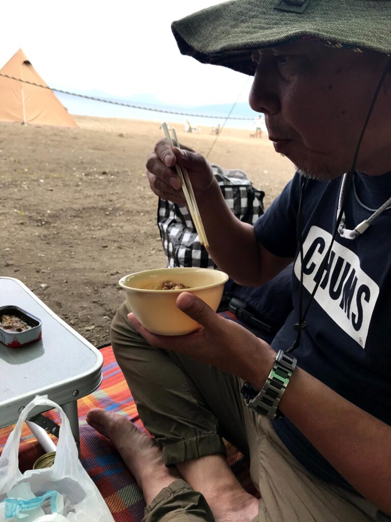 ハイエースのそばでキャンプ飯を食べる男性のアップ（支笏湖畔）