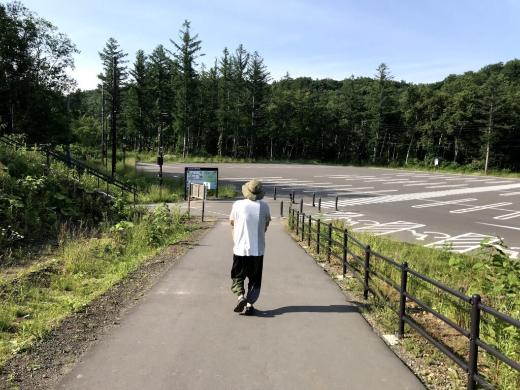 舗装された遊歩道を歩く男性と、両側に広がる草木、奥には広い駐車場が見える